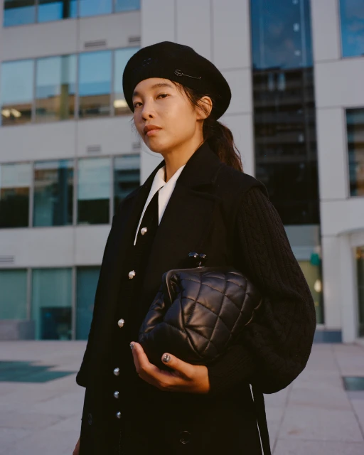 Stylish woman in a beret and coat holds a quilted leather purse in front of a modern building with glass windows, outdoors.