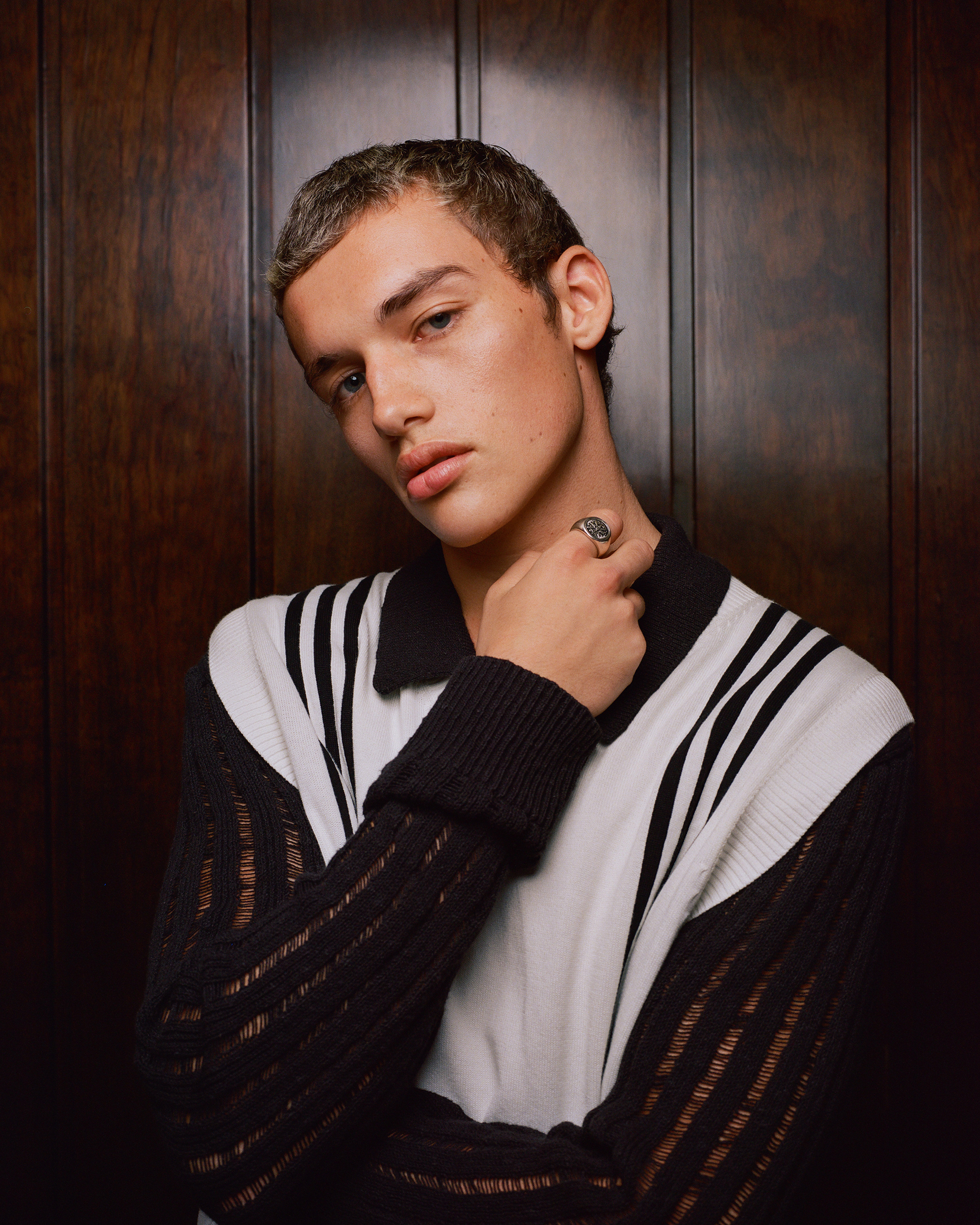Portrait of a young adult man with short, dyed hair, wearing a polo shirt and ring, posing against a dark wood paneled background.