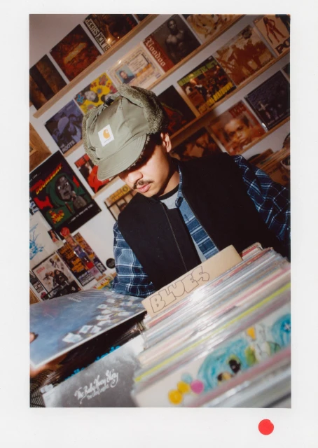 Man wearing a Carhartt hat and vest, surrounded by stacks of vinyl records and record covers in a shop with warm lighting.