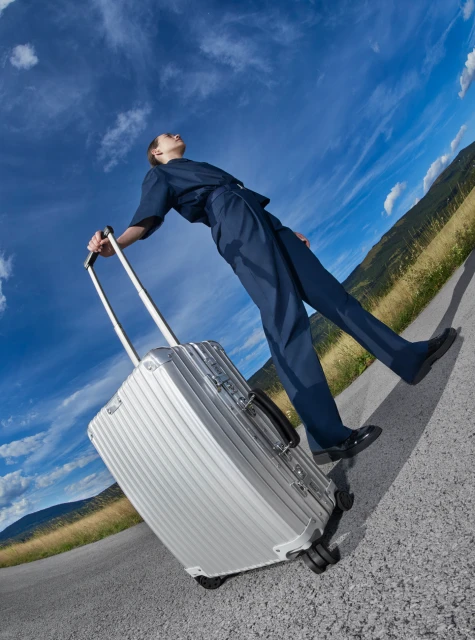 Woman pulling silver luggage on a road outdoors against a blue sky with clouds, heading toward distant mountains. Wide angle shot.