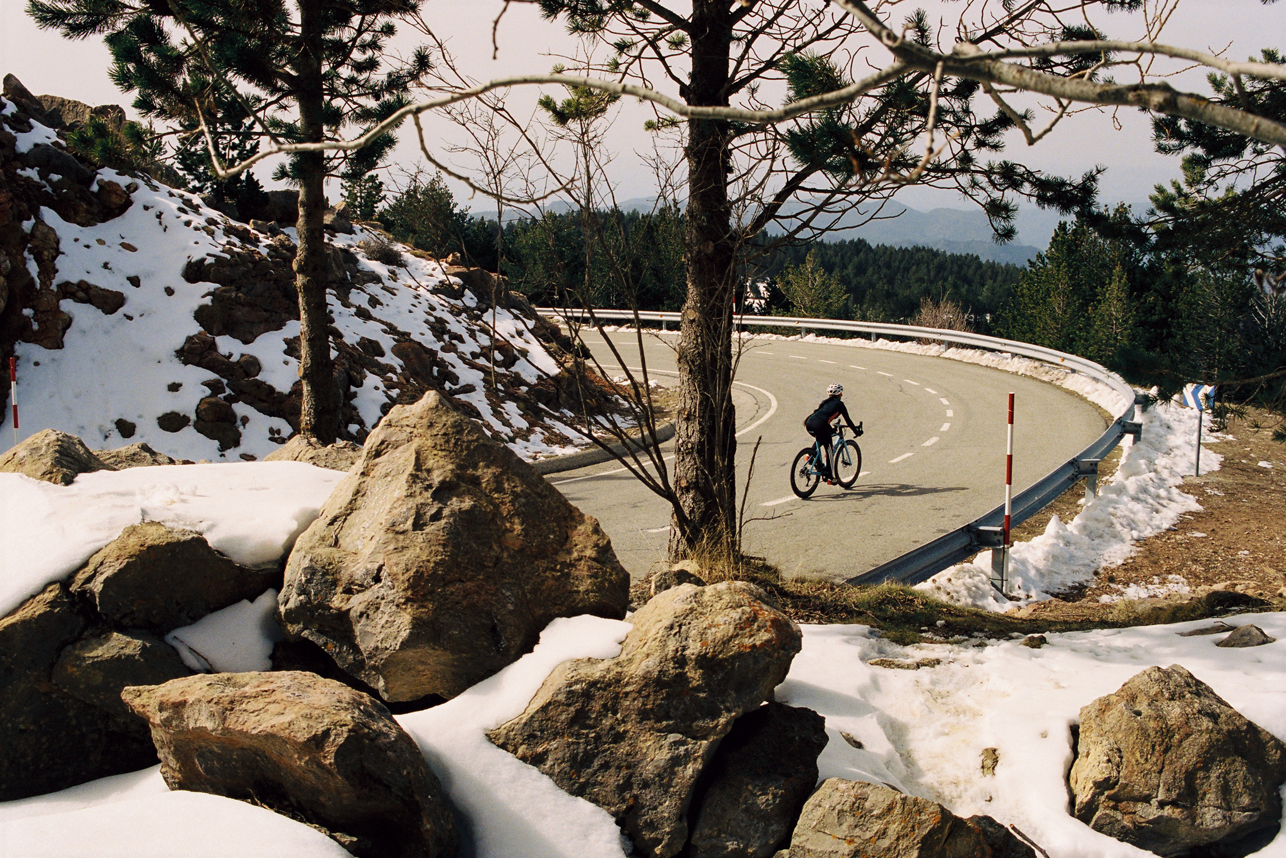 Cyclist rides on a winding mountain road surrounded by snow and trees, enjoying a winter cycling scene with a clear sky.