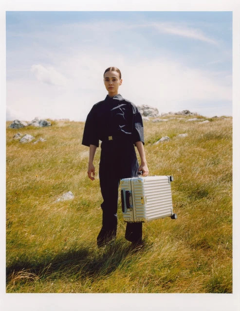 A woman in a dark blue jumpsuit stands in a grassy field with a silver suitcase under a blue sky with light clouds, shot outdoors.