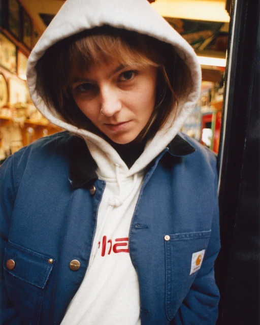 Portrait of a young adult woman with blonde hair in a hooded sweatshirt and blue jacket, standing indoors with blurry background.