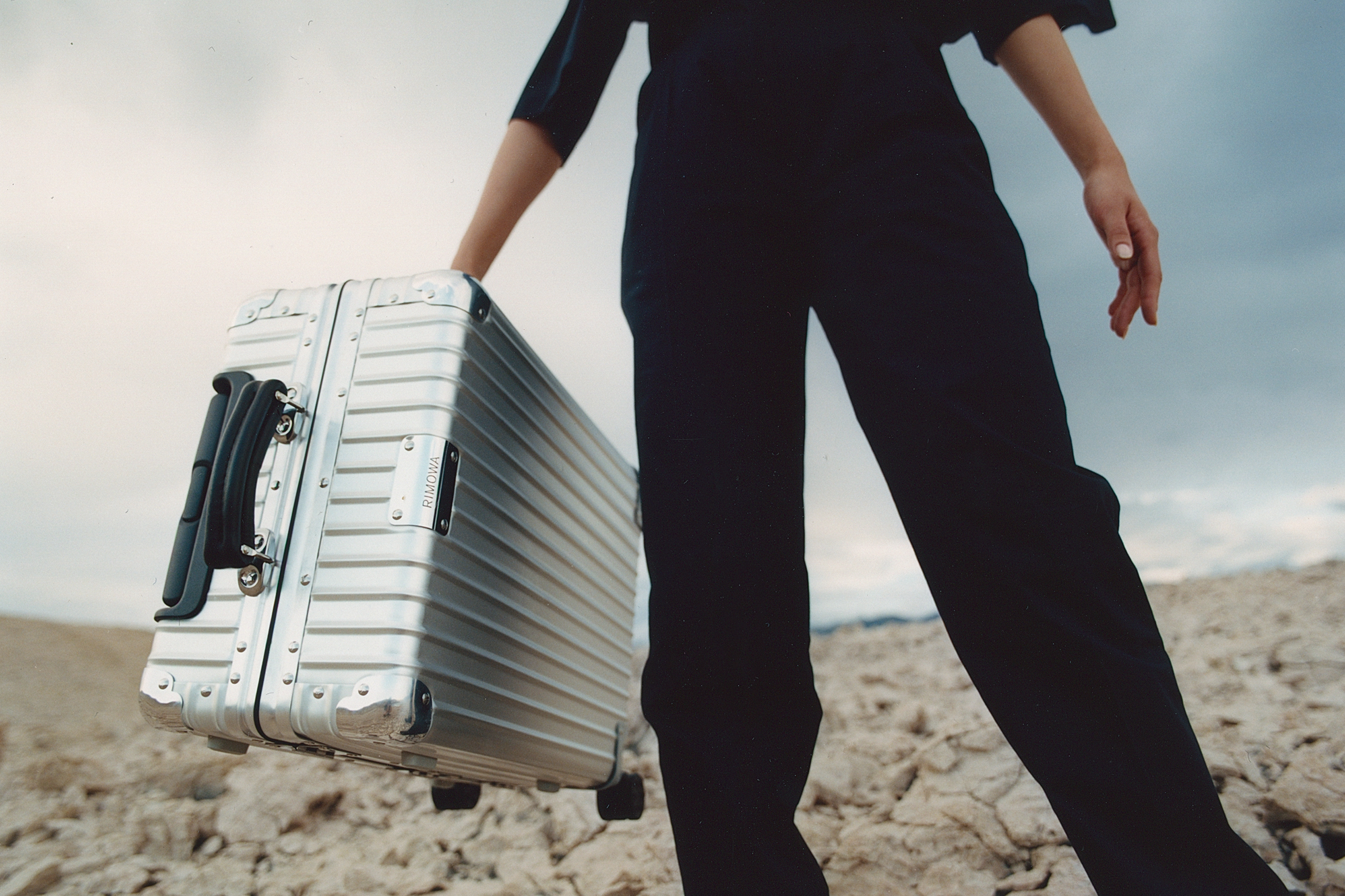 Person in dark clothing poses with a silver suitcase in a desert environment under a cloudy sky, promoting travel and style.