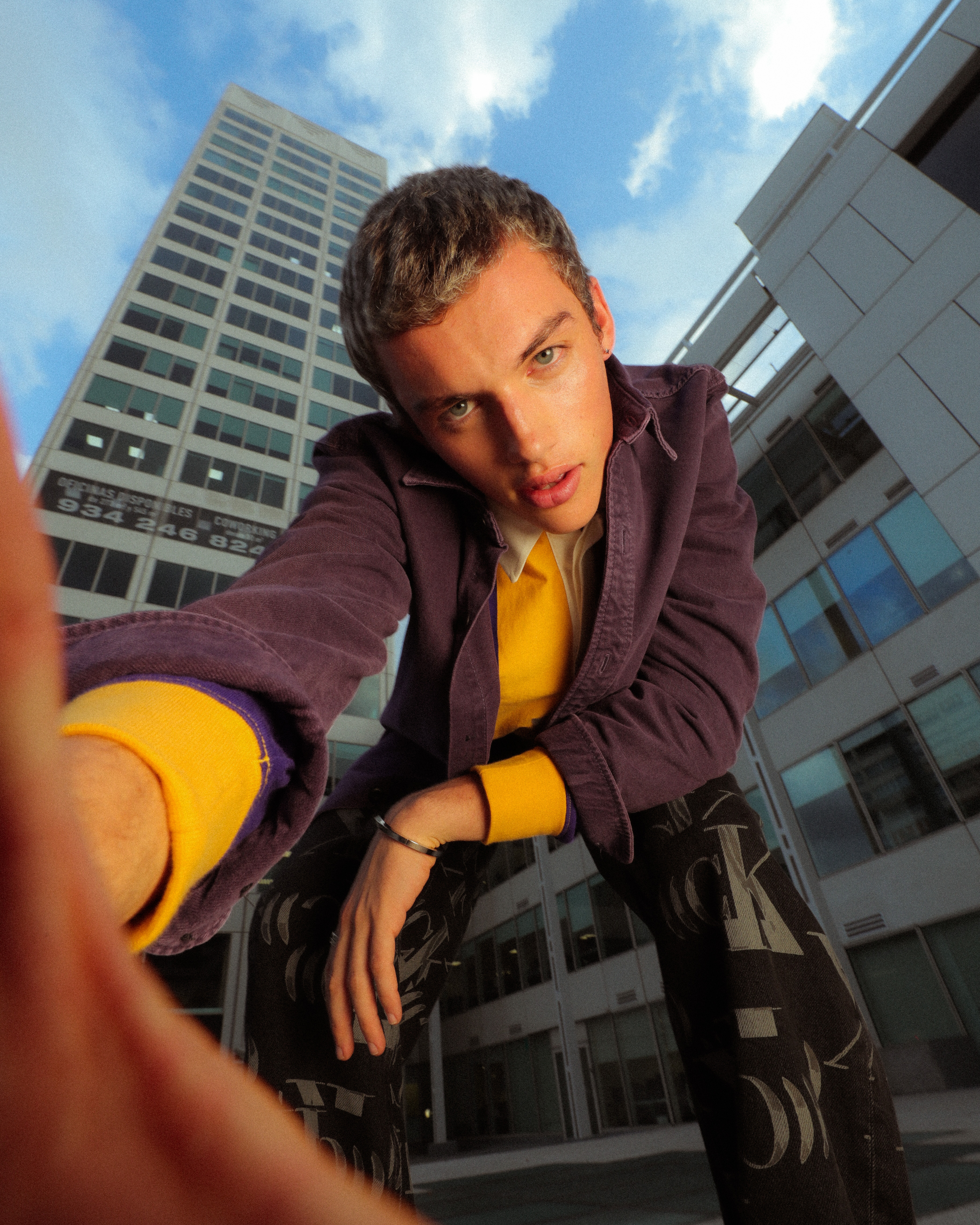 Low-angle shot of a young man in a purple jacket, yellow shirt, and patterned pants against a backdrop of blue sky and tall buildings.