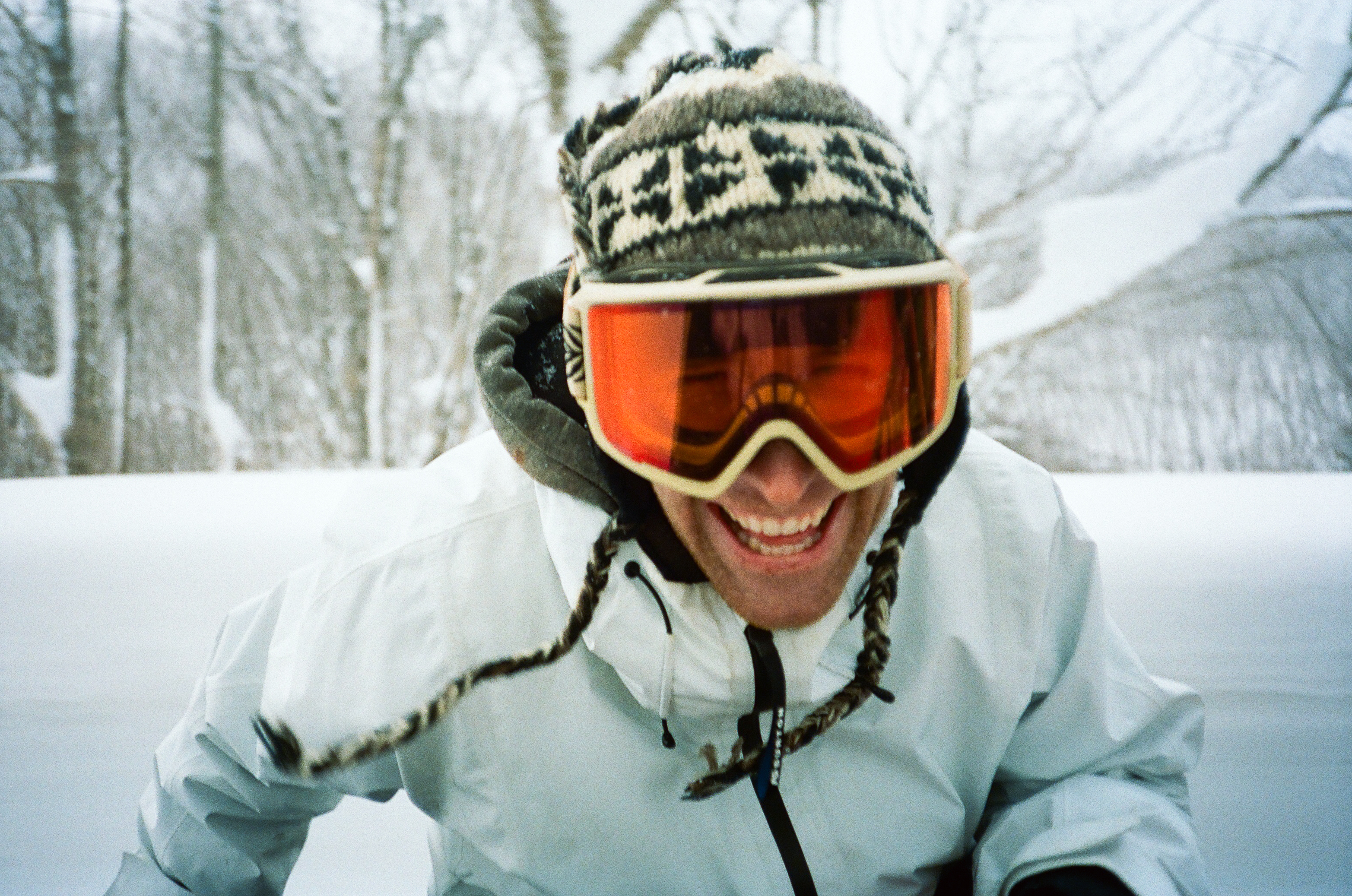 Close-up of smiling man with orange ski goggles, winter hat, and white jacket in snowy outdoor scene with blurred trees in the background.