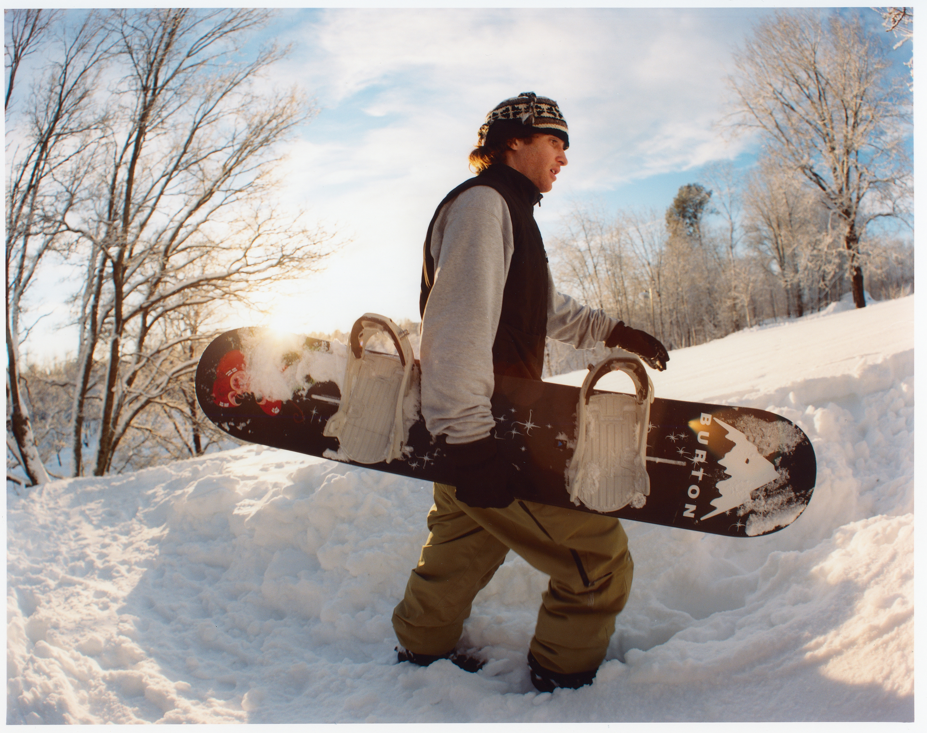 Man carries a snowboard while walking through a snowy landscape under a blue and white sky, trees lining the horizon in winter.
