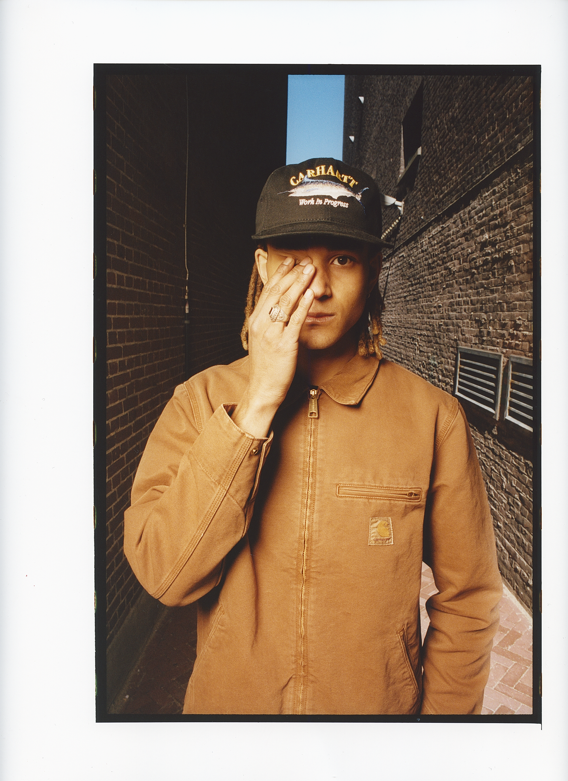 Young adult in a Carhartt hat and jacket, standing in an alley with brick walls, partially covers their face with their hand.