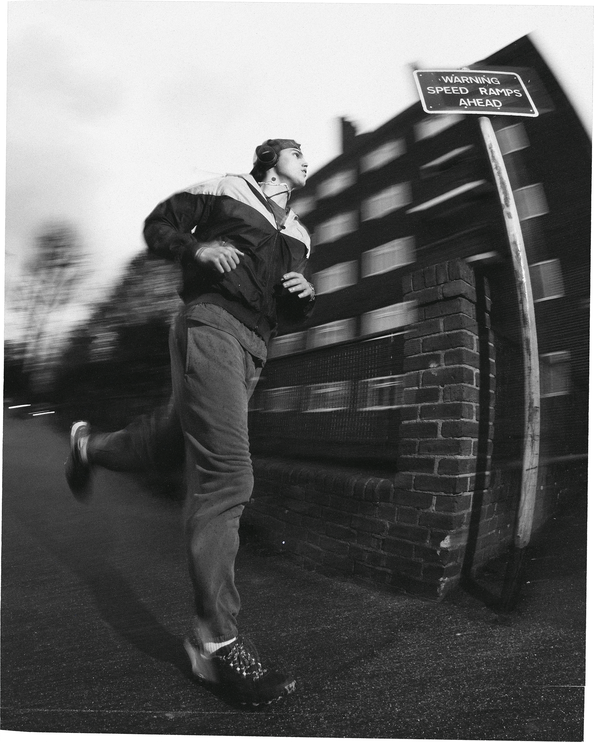 A person is seen running next to a brick building, captured in black and white with motion blur, with a 'Warning Speed Ramps Ahead' sign.