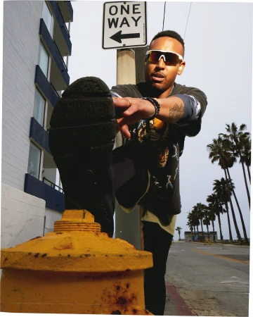 Man stretching leg on urban street with palm trees in the background. Wearing sunglasses and casual clothes, leaning on yellow bollard.