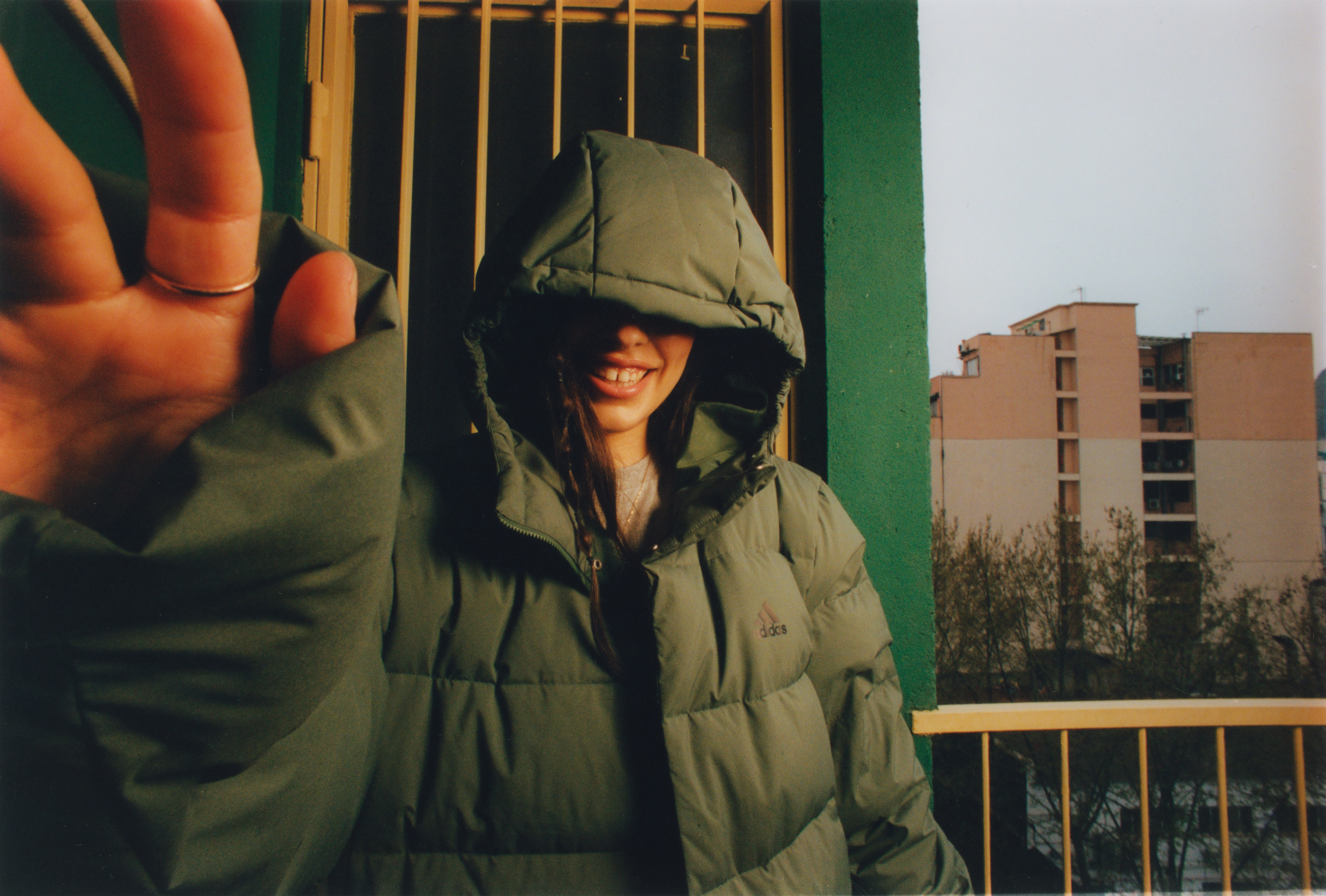 A smiling woman in a puffy green jacket with the hood up poses with her hand extended toward the camera in front of a gated balcony.