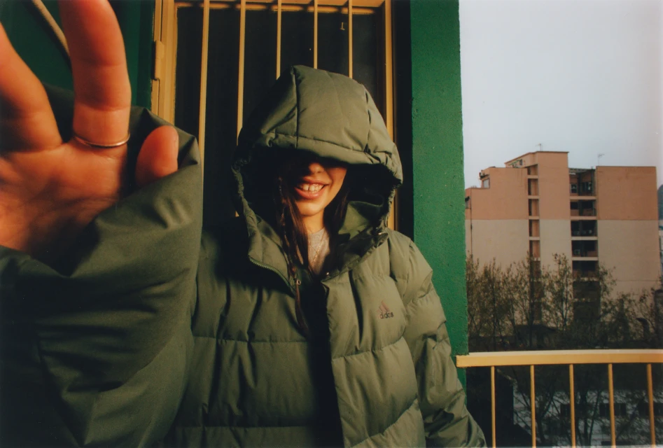 A smiling woman in a puffy green jacket with the hood up poses with her hand extended toward the camera in front of a gated balcony.