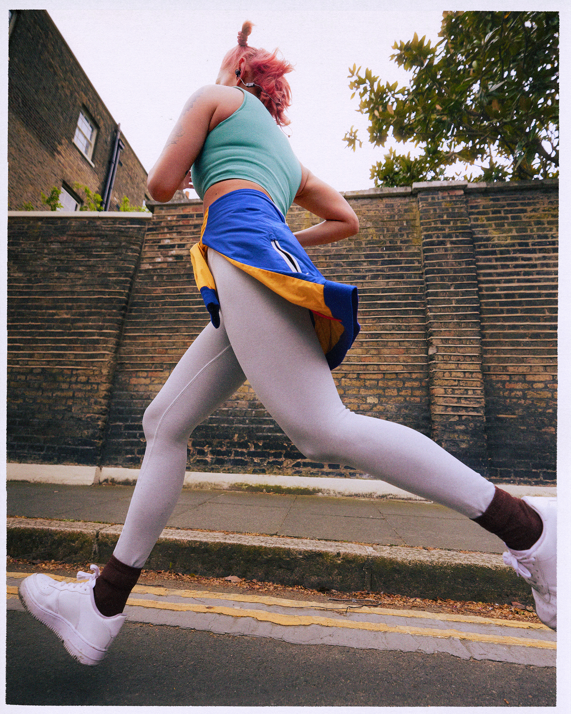 Athletic woman with pink hair in running clothes runs on a city street past a brick wall on a cloudy day, demonstrating outdoor fitness.