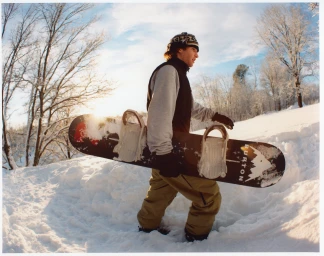 Man walking through snow carrying a snowboard under his arm, wearing a beanie, vest, and tan trousers against a snowy winter background.