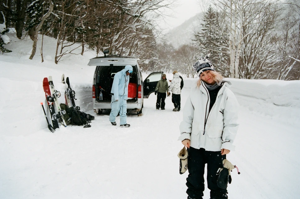 Woman in white snow jacket and beanie standing on a snowy road near a van with skis leaning against it, photographed by Arthur Couvat.