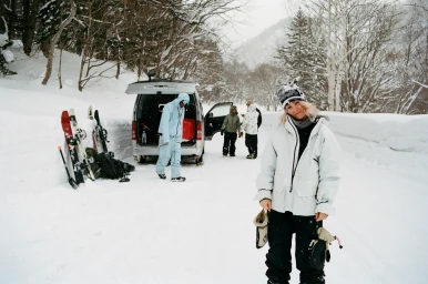 Woman in white snow jacket and beanie standing on a snowy road near a van with skis leaning against it, photographed by Arthur Couvat.