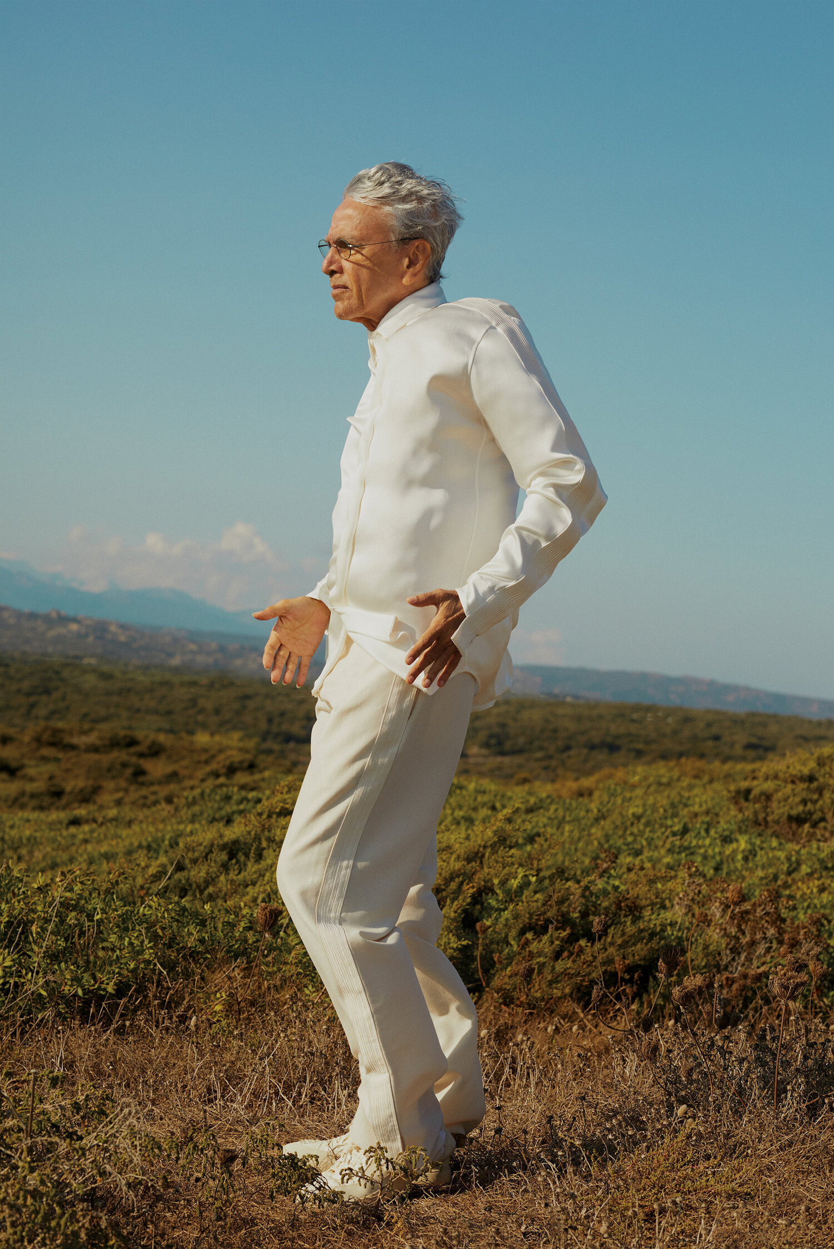 Elderly man in white suit stands in an outdoor field with mountains and blue sky in the background, looking at something out of frame.