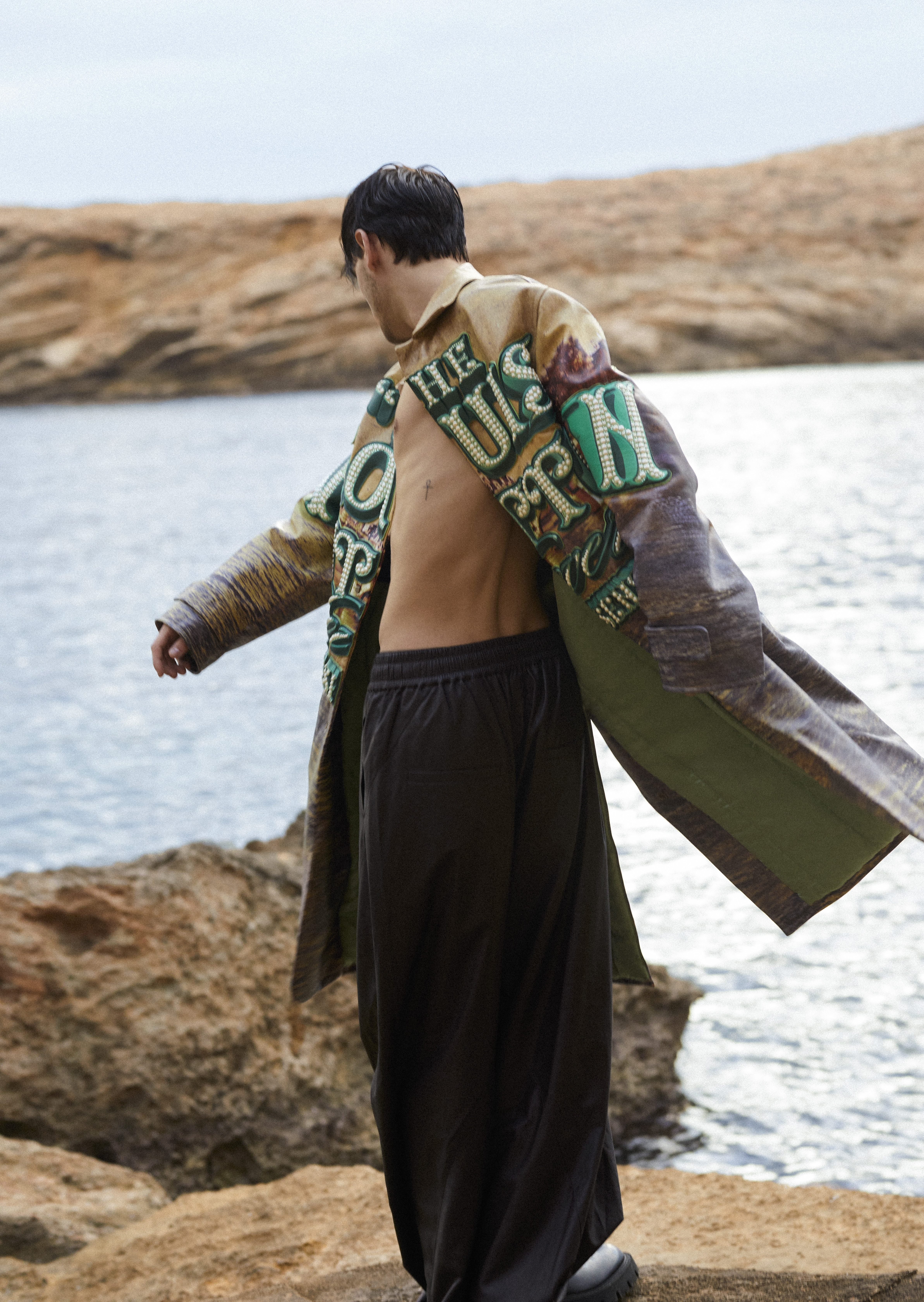 Young man on coastal rocks wearing a long, printed jacket and pants against a backdrop of water and rocky coastline.