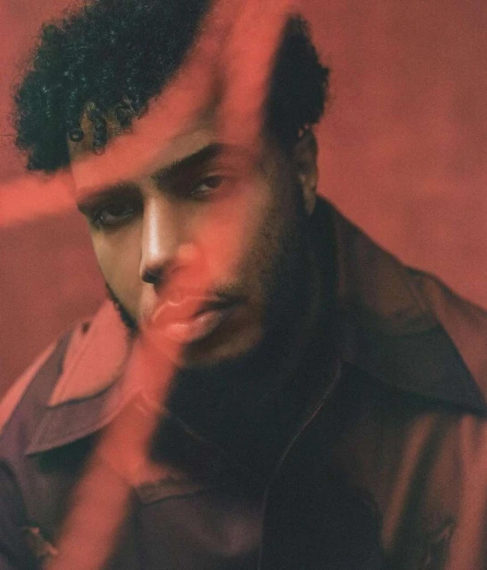 Portrait of a curly-haired adult man with brown hair and a jacket, posing in front of a red-toned background with a vertical light streak.