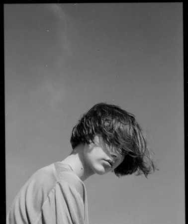 Monochrome portrait of an adult with shaggy hair, gazing slightly downward against a clear sky backdrop. Shot in natural light.