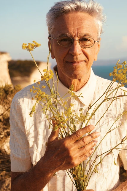 Elderly man holding yellow flowers against a scenic coastal backdrop with clear sky and textured white collared shirt in soft lighting.