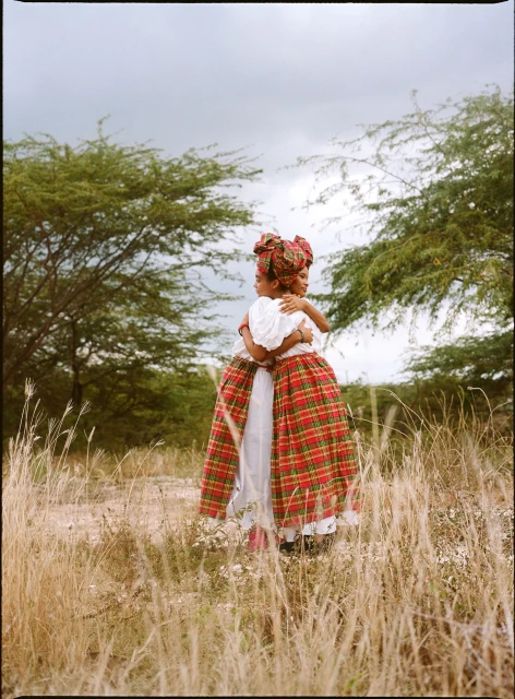 Two children embrace outdoors in patterned dresses and headwraps, set against a background of tall grass, trees, and an overcast sky.