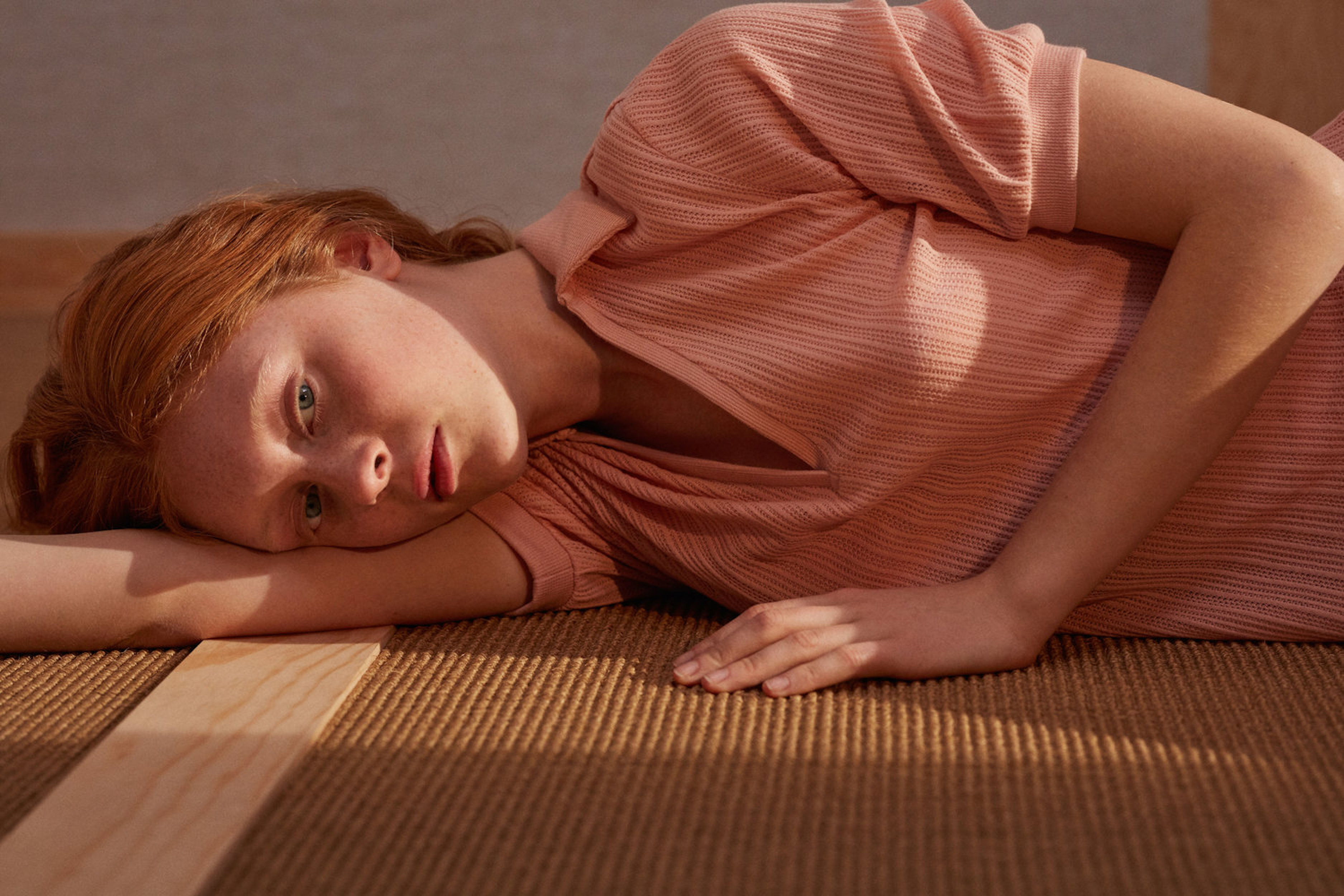 Red-haired model in a peach dress lays on a woven rug in a close-up shot with soft, diffused lighting and a warm, inviting colour palette.