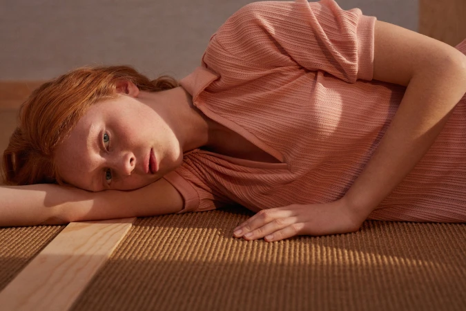 Red-haired model in a peach dress lays on a woven rug in a close-up shot with soft, diffused lighting and a warm, inviting colour palette.