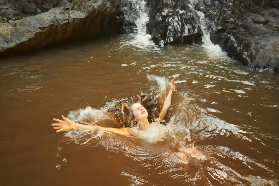 Woman in brown water with arms outstretched, surrounded by splashes, with a rocky waterfall in the background on a sunny day.