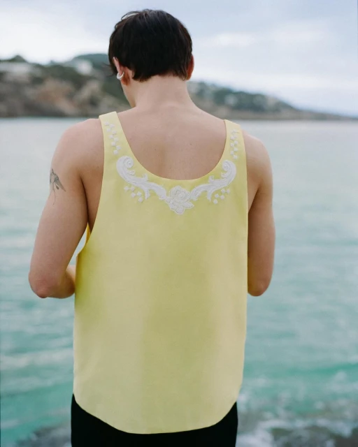 A person stands facing away from the camera wearing a yellow sleeveless shirt with white embroidery near water and land with trees.