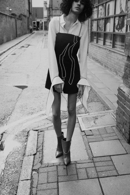 Black-and-white fashion shot of a young woman in a black mini dress and white shirt, standing in a narrow city street with brick buildings.