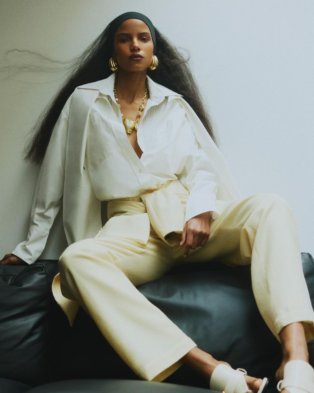 Elegant woman in a relaxed pose wearing a white blouse, light yellow trousers, gold jewelry, and a black headscarf in a studio setting.