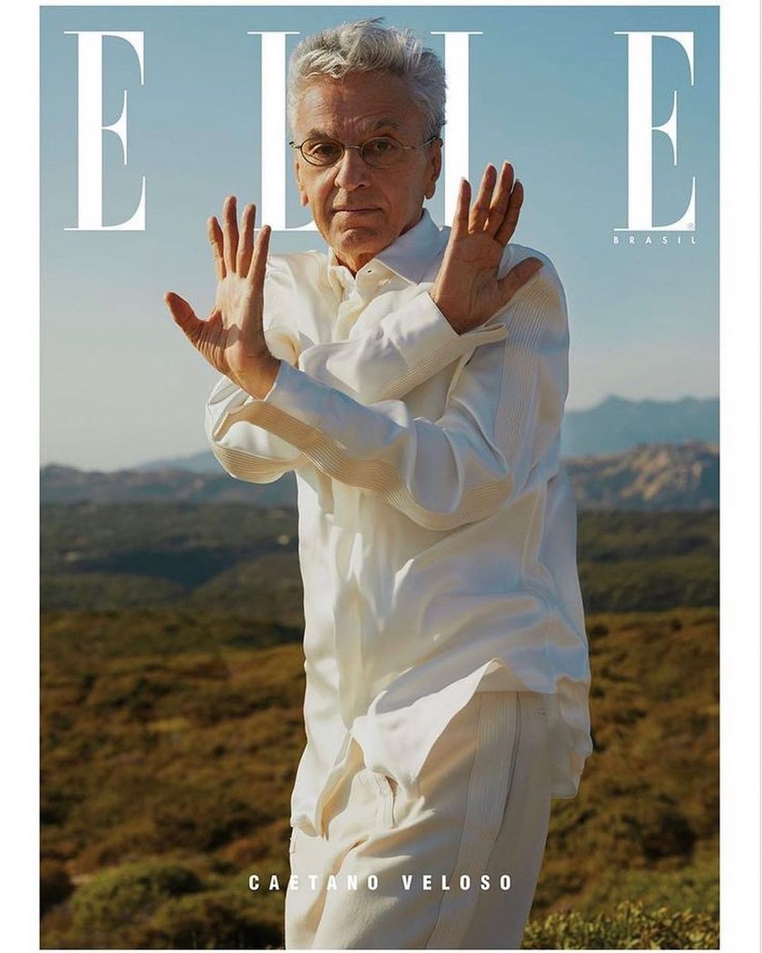 Fashion magazine cover featuring a mature male model with white hair wearing white, posing outdoors with hand gestures and a hillside background.