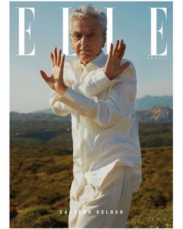 Fashion magazine cover featuring a mature male model with white hair wearing white, posing outdoors with hand gestures and a hillside background.