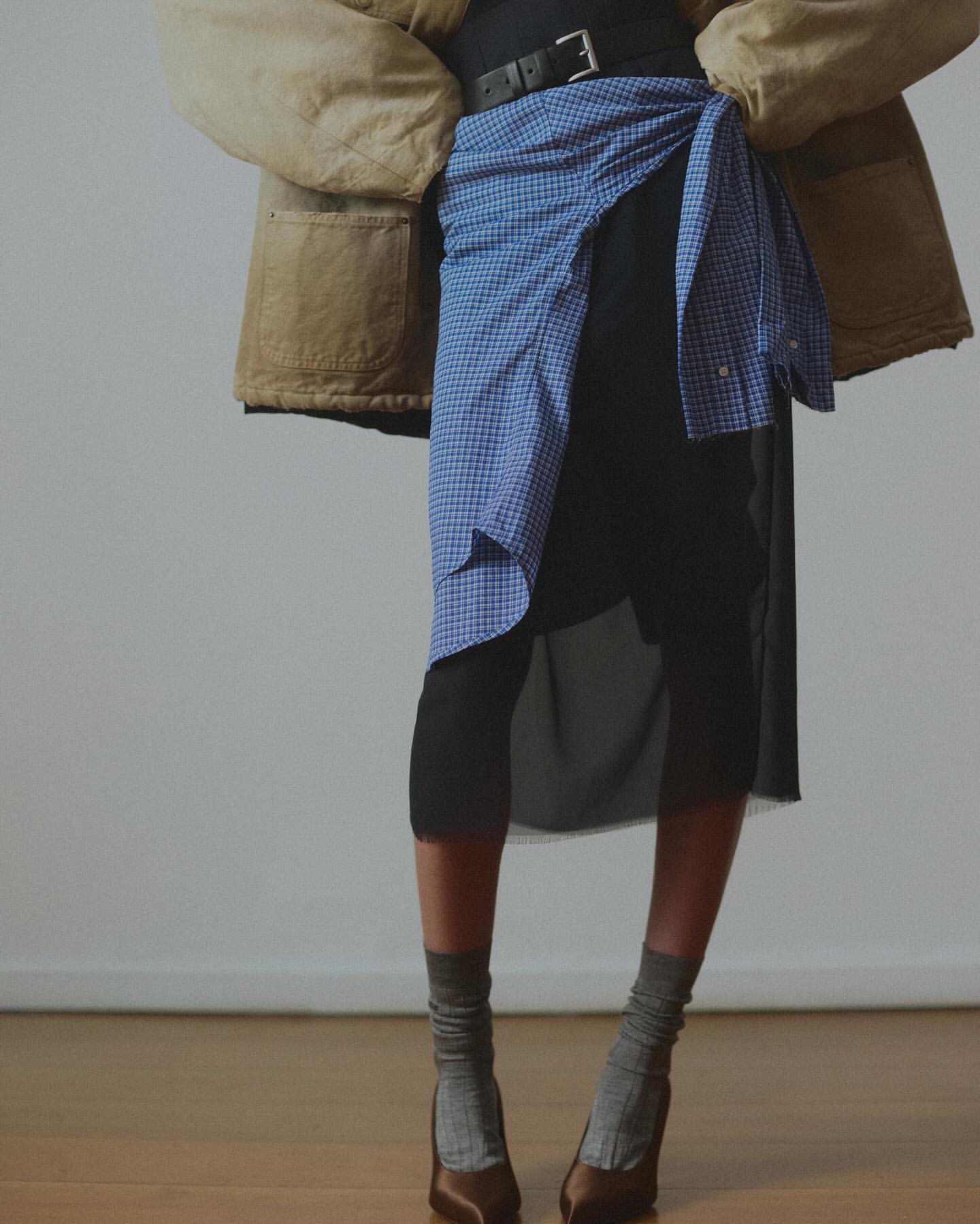 Woman wearing a brown jacket and black skirt is cinched with a blue gingham shirt, completing the look with grey socks and brown heels.