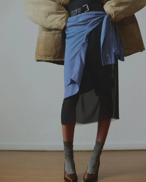 Woman wearing a brown jacket and black skirt is cinched with a blue gingham shirt, completing the look with grey socks and brown heels.