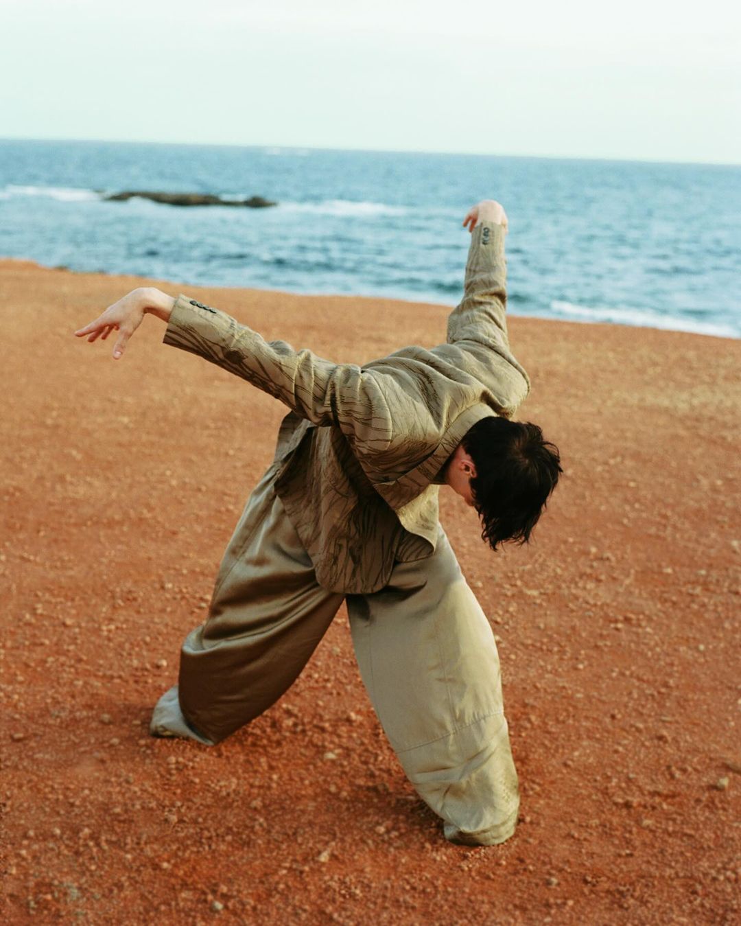 Adult woman in oversized khaki suit poses dynamically on a reddish-tan dirt surface with a blue ocean and light sky in the background.