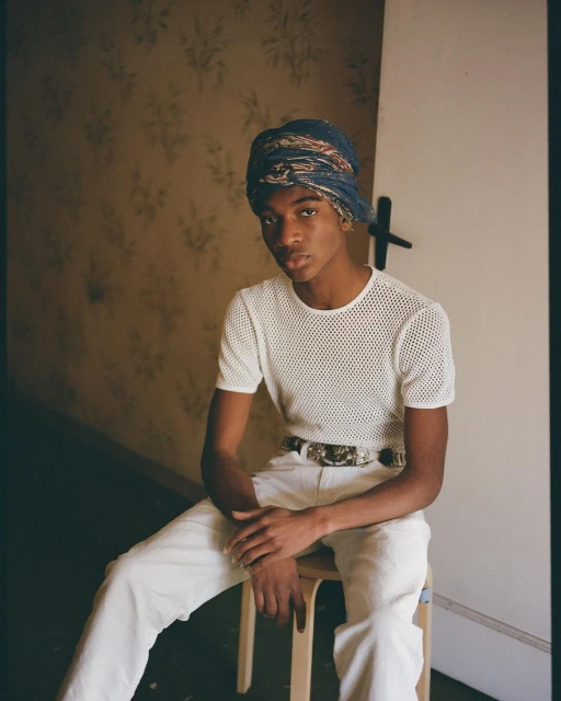 Portrait of a young adult male wearing a blue patterned head wrap, white mesh top, and white pants sitting on a wooden stool.