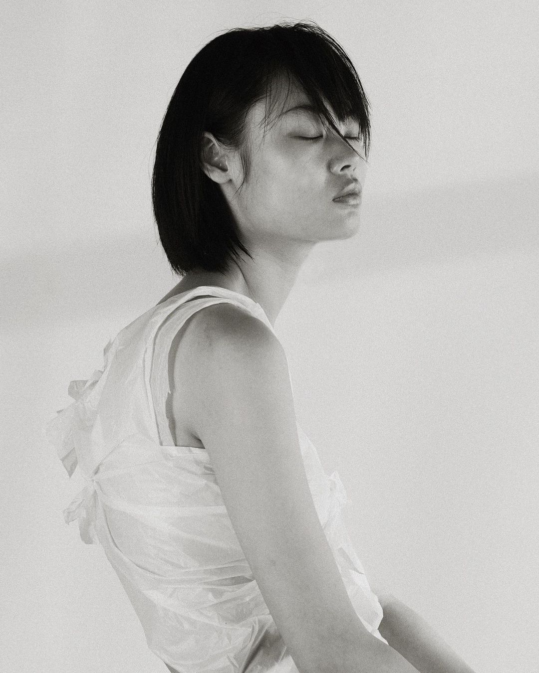 Monochrome portrait of a young Asian woman with closed eyes wearing a light-colored top against a bright background, captured in a studio.