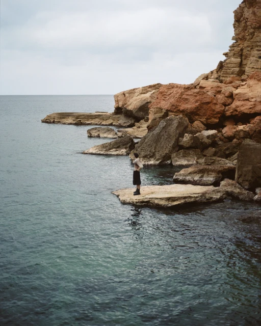 Person standing on rock formation by the sea, featuring rocky coastline, blue-green water, and a cloudy sky in a natural setting.