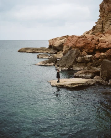 Person standing on rock formation by the sea, featuring rocky coastline, blue-green water, and a cloudy sky in a natural setting.