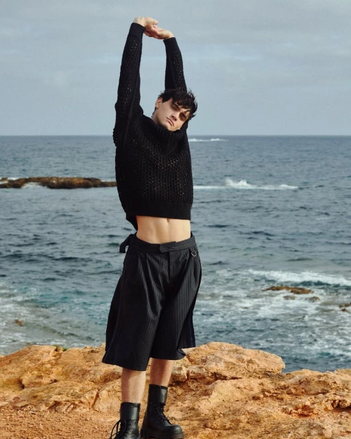 Fit adult male with dark hair and black outfit stretches on a rocky beach with ocean backdrop and cloudy skies on a sunny day.