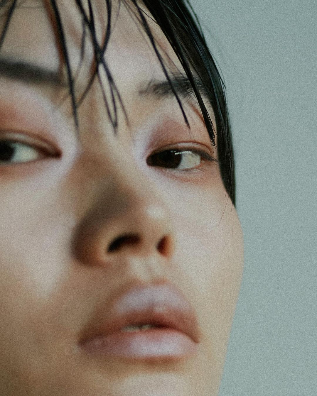 Close-up portrait of a person with wet black hair, light skin, visible makeup around the eyes, and a neutral facial expression against a grey background.