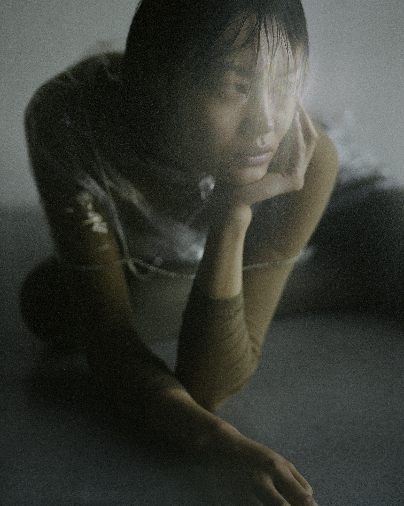 Portrait of an Asian woman with short, wet hair kneeling and resting her chin on her hand, wearing a shiny translucent top in a light studio.