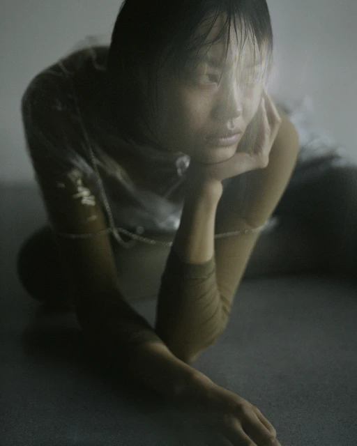 Portrait of an Asian woman with short, wet hair kneeling and resting her chin on her hand, wearing a shiny translucent top in a light studio.