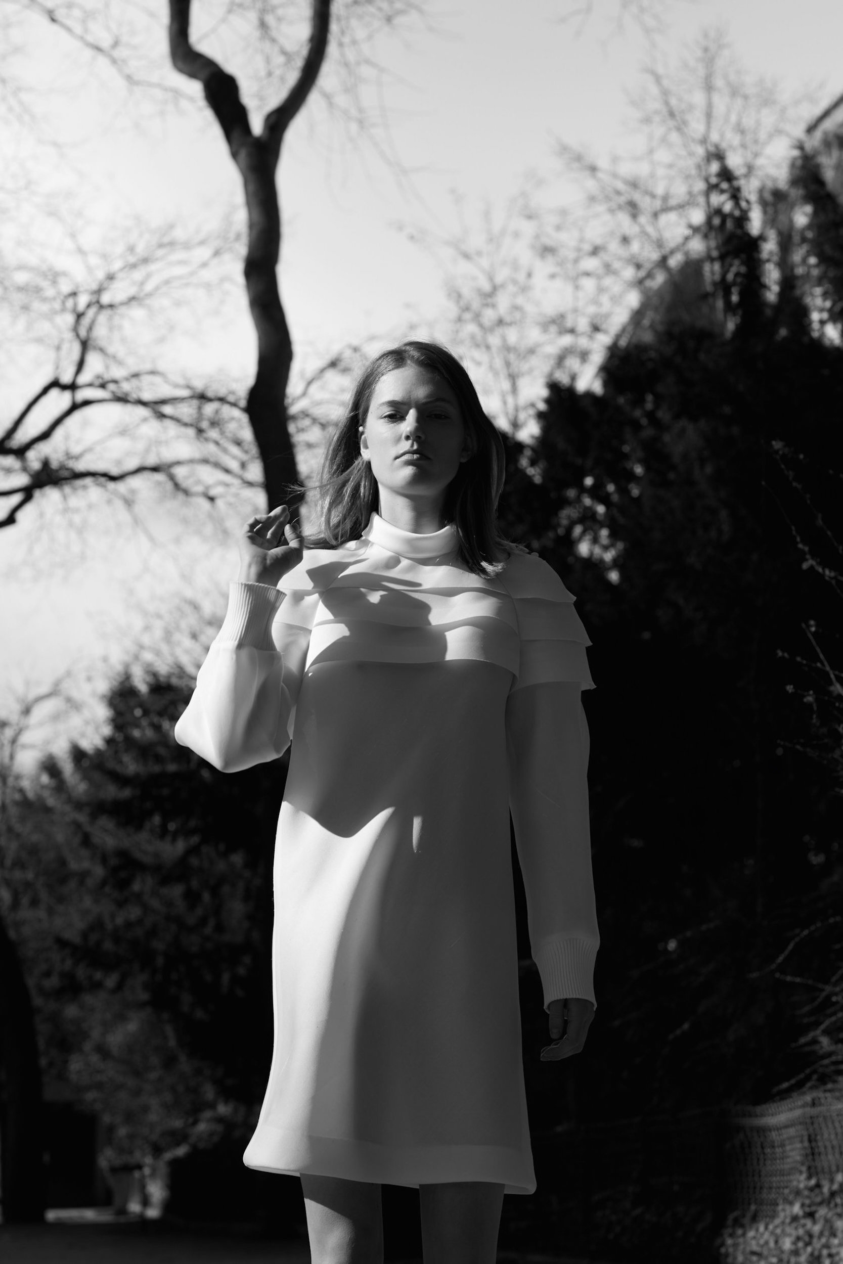 Monochrome portrait of a young fair-skinned woman in a white dress standing outdoors against a backdrop of trees and a building.