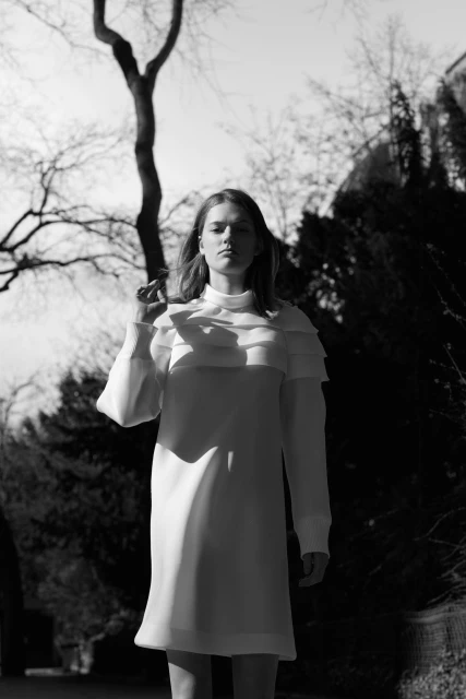 Monochrome portrait of a young fair-skinned woman in a white dress standing outdoors against a backdrop of trees and a building.