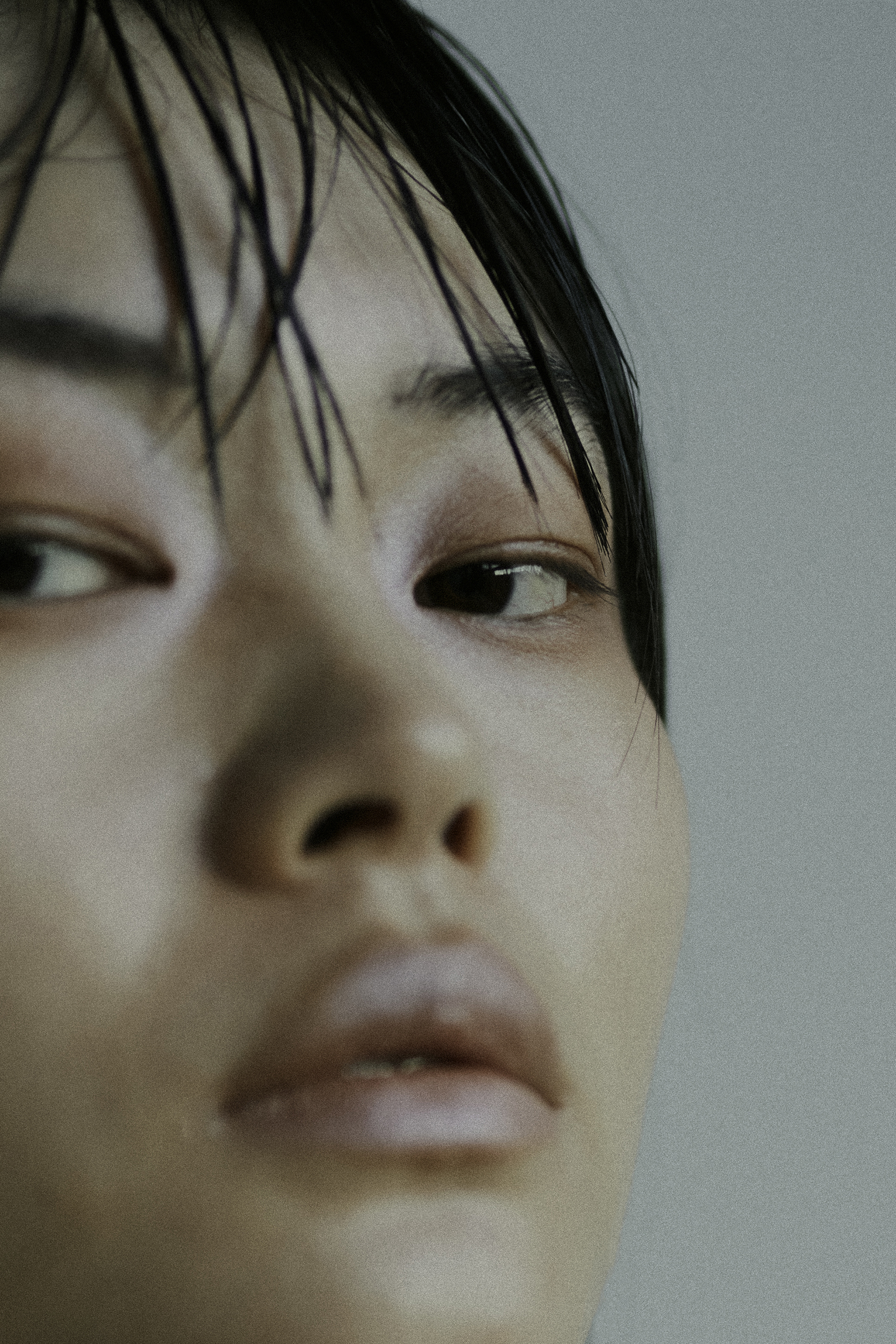 Close-up portrait of an East Asian woman with wet dark hair and neutral makeup against a plain grey background