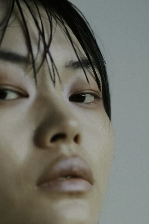 Close-up portrait of an East Asian woman with wet dark hair and neutral makeup against a plain grey background