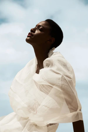 Black woman with short hair wearing sheer white draped fabric looking up against a cloudy sky
